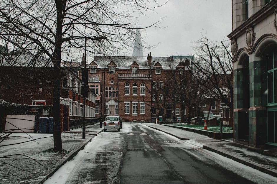 A narrow residential street during winter with snow covering the pavement and some snow settled on the rooftops of brick and stone buildings. The street is lined with leafless trees on both sides, their branches extending overhead. On the left, there are a few houses with bay windows and small front gardens, partially obscured by snow, while on the right, a larger building with decorative stonework and arched windows is visible. A parked silver car is situated near the middle of the street, close to the curb. The background features a prominent red-brick building with multiple chimneys and turrets, suggesting an older architectural style. The scene appears overcast, with diffused daylight creating a muted, greyish atmosphere. This setting exemplifies a typical urban environment where independent rubbish collection services like Rubbish Clearance Bermondsey might handle on-site waste disposal, especially during winter months when street cleanliness and waste removal are crucial for maintaining a tidy appearance in residential areas.