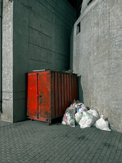 A narrow, urban alleyway cluttered with discarded rubbish and debris, including a large grey fabric sack with visible seams and a metal handle, positioned at the forefront on the uneven, dark asphalt ground. In the background, there are overflowing piles of cardboard boxes, plastic bags, and miscellaneous waste stacked haphazardly against the back wall, which appears weathered and partly covered in graffiti. The alley is flanked by tall buildings on both sides, one with a brick facade and the other painted in muted tones, featuring small windows and metal security bars. Sparse, leafless tree branches extend overhead, hinting at an urban environment during colder months. Natural, diffused lighting provides a subdued atmosphere, emphasizing the cluttered scene typical of private waste accumulation in an alley used for rubbish collection. This scene suggests a location where independent waste clearance, possibly by companies like Rubbish Clearance Bermondsey, is utilized for managing overflowing waste outside regular municipal collection services.
