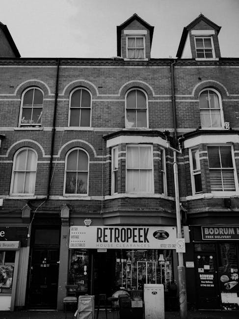 The image depicts the facade of a multi-storey brick building with several large arched windows on the upper floors, typical of Victorian architectural style. The ground floor features a retail unit with a sign reading 'RETROPEEK HOUSE CLEARANCES' in bold, contrasting lettering, indicating a business specializing in waste clearance or junk removal services. In front of the shop, there are various items, including a small table and some containers, suggesting recent activity related to rubbish collection or clearance. To the right, another storefront with signage is partially visible, offering delivery services. The building is situated directly on a paved street or sidewalk with no visible vehicle for clearance or waste disposal in the immediate vicinity. The overall environment appears to be in an urban setting, and the monochrome tone emphasizes the textures of the brickwork and the orderly arrangement of the shopfronts. This scene subtly reflects the local commercial activity linked to private waste handling, supporting the services provided by Rubbish Clearance Bermondsey for independent rubbish removal efforts.