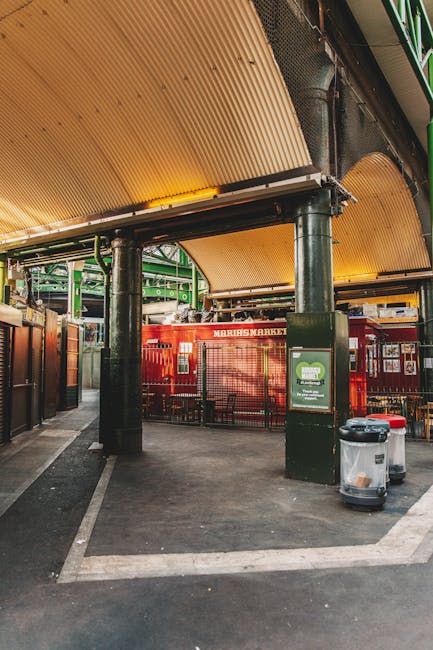 The image depicts an indoor market entrance with a high, curved, ribbed wooden ceiling supported by dark green metal columns. In the foreground, there are two large waste bins, one with a transparent lid and the other with a red lid, positioned on a paved surface near a black post, which may contain informational signage. Behind the bins, a red metal railing separates the walkway from the market area, which includes a red and white sign indicating 'Marius Market' on the building facade. The surrounding environment appears clean and organized, with natural lighting illuminating the space, highlighting the textures of the wooden ceiling and the glossy finish of the metal structures. The scene suggests a setting where appliances or debris may be cleared regularly, aligning with private waste management and rubbish removal services such as those provided by Rubbish Clearance Bermondsey, particularly for urban sites like markets or commercial premises that may require scheduled or independent rubbish clearance solutions.