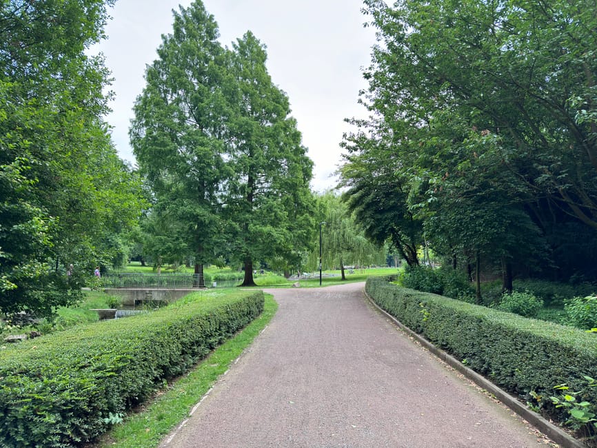 A wide, paved pathway in a park surrounded by lush greenery, including neatly trimmed hedges along both sides and tall trees with dense foliage overhead. The path gently curves to the right in the distance, leading towards open grassy areas and several lampposts. On the left side, a small water feature with a concrete edge is visible among the trees, and the environment appears calm and well-maintained. The scene is illuminated by natural daylight with a partly cloudy sky, creating a soft, diffuse light that highlights the vibrant green tones of the leaves and grass. This outdoor setting, featuring the tidy park landscape, relates to private or independent waste management, as Rubbish Clearance Bermondsey occasionally supports local rubbish removal within community parks and public spaces, ensuring cleanliness and upkeep of outdoor environments in Bermondsey.SE16.