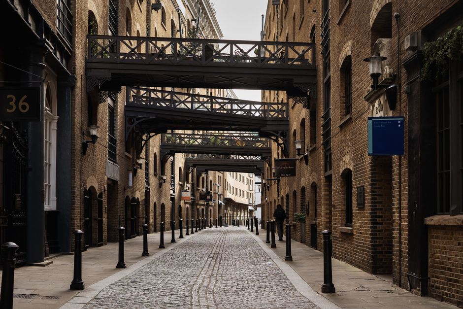 A narrow cobbled street lined with brick buildings on both sides, featuring multiple black metal pedestrian bollards along the edge of the pavement. The buildings have a historic appearance with arched windows, some of which are accented with black wrought iron details. Above the street, there are several decorative black metal footbridges connecting the upper floors of the buildings, extending across the alleyway. The scene is illuminated with natural daylight, casting soft shadows and highlighting the texture of the cobblestones and brickwork. A person dressed in dark clothing walks away from the camera towards the distant end of the street. The overall environment appears urban with a mixture of modern and historic architectural elements, typical of a city area that may include private or independent property management, supporting alternative waste disposal options in a residential or commercial setting relevant to rubbish removal services.