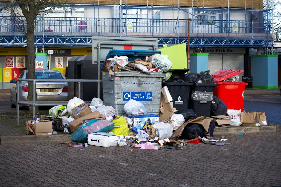 A collection of overflowing rubbish scattered on a paved area beside a parked silver vehicle, with various black, red, and brown waste bags, flattened cardboard boxes, and loose paper cluttering the space. The black waste bags appear full and tied, while the cardboard boxes are flattened or partially torn, with some spilling contents onto the ground. A large gray metal recycling bin labeled for mixed paper and cardboard is positioned in the center, filled with paper and packaging materials, with some waste protruding from its open lid. To the right, a black bin for general waste and a red bin for recyclable materials are placed nearby, both filled and with waste bags leaning against them. In the background, a building with blue scaffolding and a yellow storefront with signage can be seen, along with a tree on the left side. The scene is outdoors, on an urban pavement, suggesting a situation where independent rubbish clearance or collection services, such as those from Rubbish Clearance Bermondsey, might be involved in handling the waste segregated outside an office or commercial premise.