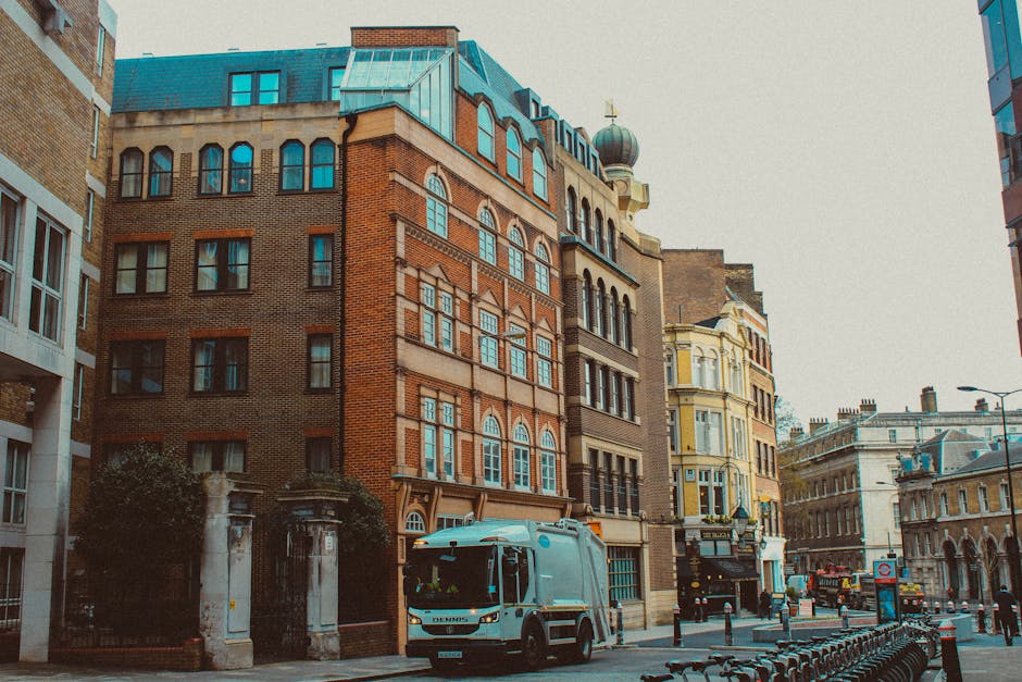 A street scene featuring a row of historic multi-story buildings with diverse architectural details, including arched and rectangular windows, brick facades in shades of red and beige, some with decorative stone accents, and a dome-topped tower in the background. In the foreground, a white rubbish collection vehicle with blue markings is parked alongside a sidewalk, partially blocking the pedestrian walkway. The vehicle, associated with independent waste collection services like Rubbish Clearance Bermondsey, is positioned near an entrance surrounded by dark wrought-iron gates and stone pillars, suggesting a private or commercial property. To the right, there are street lampposts and a line of bicycles secured to a bike rack, with a clear daytime sky providing even lighting that highlights the textures of the stone, brick, and glass surfaces. The scene reflects typical urban rubbish removal operations close to business and residential premises, emphasizing the utilitarian aspect of on-site clearance and private waste handling in a historic city setting.